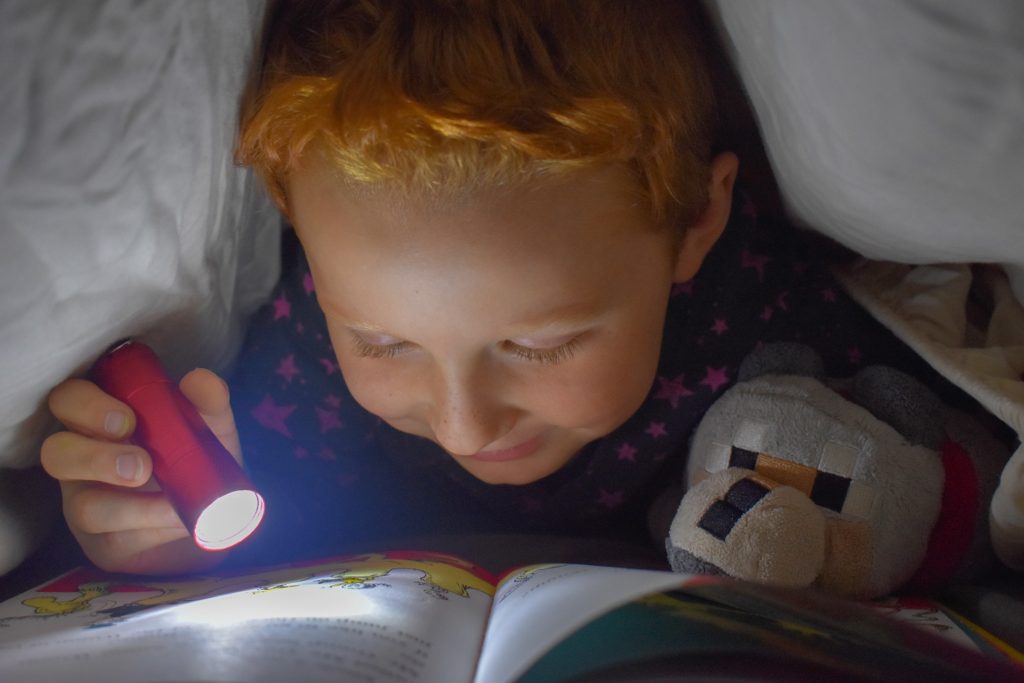 Picture of a boy learning by reading a book using a flashlight under his covers